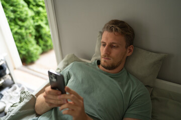 a young handsome man in a green t shirt lies in bed and stares at his phone