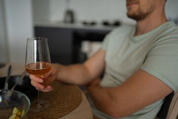 A young man enjoying a refreshing beverage in a glass, seated comfortably in a modern kitchen or dining area. He appears relaxed, sipping from a wine or cocktail glass, giving a casual vibe.