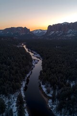 Obraz premium A serene winter landscape showcases a winding river surrounded by lush pine forests and majestic mountains at sunset, creating a tranquil and picturesque scene.