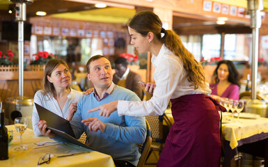 Polite young waitress using smartphone to take order in restaurant, helping couple choose dishes from menu..