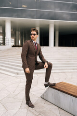 Full length vertical portrait of serious confident businessman in stylish suit and sunglasses posing outdoors, exuding success and professionalism, city architecture complementing modern urban look.