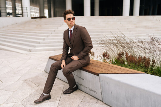 Full length portrait of of stylish photogenic young businessman in brown suit and sunglasses sitting confidently on city bench, exuding professionalism and success in modern urban setting.