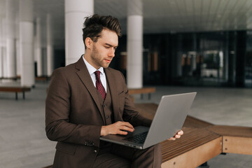 Serious focused businessman in stylish sharp brown suit working typing on laptop computer sitting outside by modern office building, showing determination and ambition in professional urban setting.