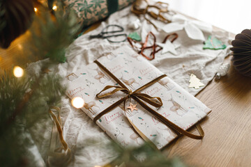 Stylish wrapped christmas gift, modern paper ornaments and pine branches on rustic table on background of christmas lights bokeh. Merry Christmas! Atmospheric christmas preparation