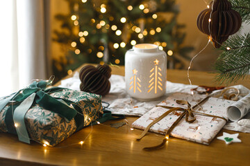 Stylish wrapped christmas gift, glowing lantern and modern paper ornaments on rustic table on background of christmas tree lights bokeh. Merry Christmas! Atmospheric christmas time