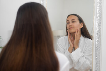 Obraz premium A woman, in her cozy bathrobe, practices a beauty skincare routine at home, studying herself in the bathroom mirror. 