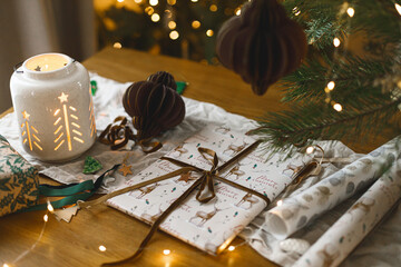 Stylish wrapped christmas gift, glowing lantern and modern paper ornaments on rustic table on background of christmas tree lights bokeh. Merry Christmas! Atmospheric christmas time