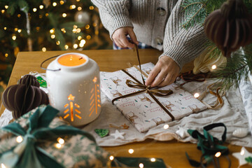 Woman wrapping stylish christmas gift on rustic table with modern paper ornaments and lantern on background of christmas tree lights bokeh. Merry Christmas! Atmospheric christmas time