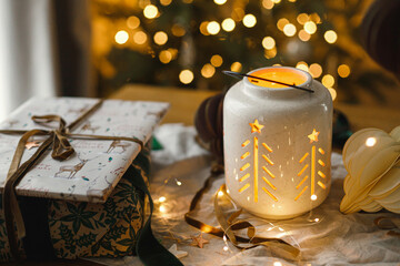 Merry Christmas and Happy holidays! Stylish christmas gift, glowing lantern, modern paper ornaments on table with golden christmas lights. Atmospheric christmas eve, holiday magical time