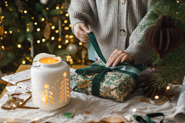 Woman wrapping stylish christmas gift on rustic table with modern paper ornaments and lantern on background of christmas tree lights bokeh. Merry Christmas! Atmospheric christmas time