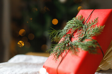 Stylish christmas red gift with modern cedar wreath on table on background of christmas tree lights bokeh. Merry Christmas! Atmospheric christmas time, holiday banner