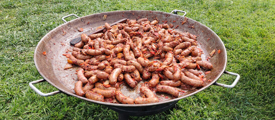 A large paella pan filled with sizzling chorizo sausages in a Pomarola tomato sauce, cooked outdoors at a party