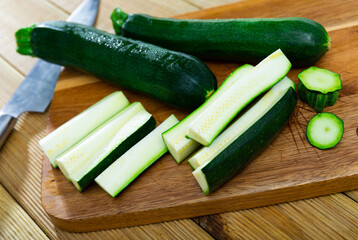 Against background of wooden table, several large pieces of zucchini are laid out on board. Ripe vegetables on chopping board before cooking dinner. Cut some zucchini fruit into oblong segments, parts