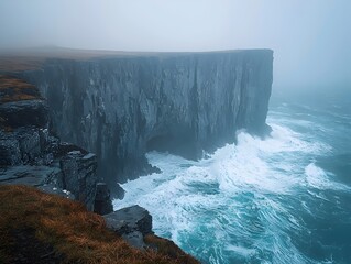 A dramatic coastal cliff in misty conditions, showcasing jagged rock formations and crashing waves below, creating a serene yet powerful atmosphere. - Powered by Adobe