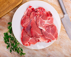 Slices of fresh raw pork loin head with fat marbling, prepared for roasting on plate, placed on table with sprigs of fresh aromatic parsley and sharp knife. Ingredients for cooking