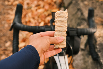 Close-up of a cyclist's hand holding a protein bar during a bike ride.  Autumn leaves and a blurred bike background create a sense of outdoor activity and healthy snacking.