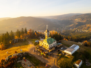 The Tanvaldsky Spicak lookout tower stands prominently in the Jizera Mountains, surrounded by vibrant autumn foliage during a serene evening glow. Visitors are seen enjoying the view.