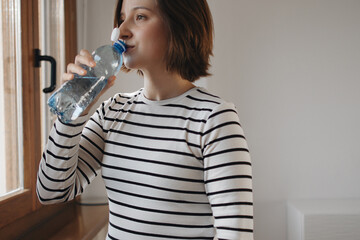 Woman Drinking Water From A PET Plastic Bottle, Demonstrating The New Tethered Caps On Plastic Water Bottles, Refreshing Herself At Home. New Eu Regulation