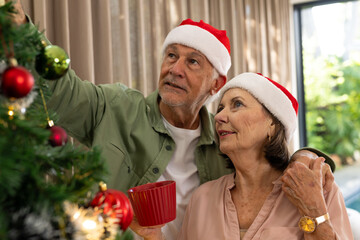 Senior couple decorating Christmas tree at home, wearing Santa hats, smiling warmly