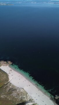 Gliding over Ons Island towards Melide Beach. Dirt path runs above the beach, surrounded by bushes. Dunes and white sand frame crystal-clear, turquoise water. Sunny Pontevedra estuary in background.