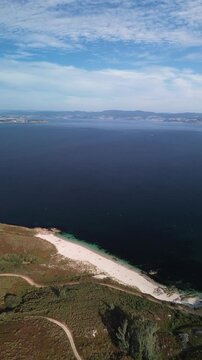 Gliding over Ons Island towards Melide Beach. Dirt paths run above the beach, surrounded by bushes. Dunes and white sand frame crystal-clear, turquoise water. Sunny Pontevedra estuary in background.