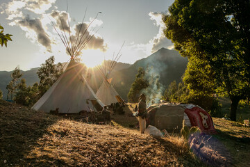 Native community with people in a landscape with teepee and temazcal in a sunrise with the sun rising over the mountains. 