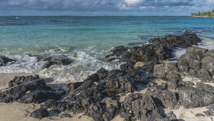 A pile of picturesque volcanic boulders on a sandy beach. Black stones on white sand are washed by the waves of the turquoise ocean. Foam. Blue sky, clouds. Mauritius. Belle Mare