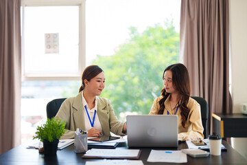 Two businesswomen in formal attire collaborate on a laptop in a bright office. They are both wearing blue lanyards and discussing ideas in a modern workspace.