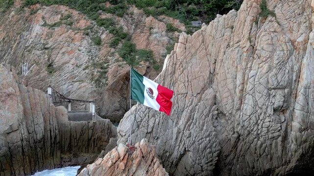Close-up aerial shot of the Mexican flag waving majestically by the rocky cliffs of La Quebrada, Acapulco, with the ocean in the background