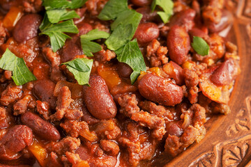 Close up view of traditional mexican spicy homemade chili con carne stew made of kidney or pinto beans, fried ground beef, tomatoes and chilli pepper served on plate decorated with fresh green parsley