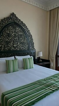 A king-sized bed with green striped pillows and a matching runner sits in a hotel room with an ornate black headboard and white curtains