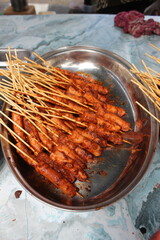 A tray filled with skewered grilled sausages coated in a spicy sauce, displayed on a metal tray at...