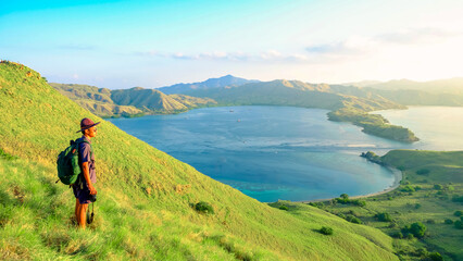 Man stands on the edge of a cliff looking at the stunning scenery with green hills and blue sea at sunrise in Gili Lawa, Komodo National Park, Indonesia. summer background and summer holiday concept. © Balnyes Visuals