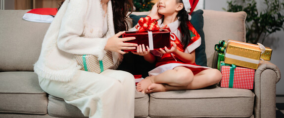 Asian mother and daughter in Santa hats enjoying a Christmas celebration with cake, gifts, and festive decor.