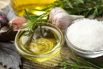 Cooking oil in bowl, salt, garlic and rosemary on wooden table, closeup
