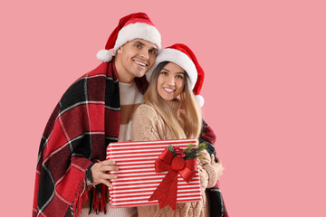 Happy young couple in Santa hats with plaid and Christmas gift on pink background