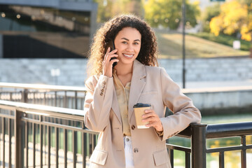 Fototapeta premium Beautiful young happy African-American businesswoman with cup of coffee talking by mobile phone near railing on embankment