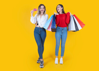 Naklejka premium Excited happy young fun two full body women wear casual clothes hold in hand credit bank card and shopping paper bags isolated on plain yellow background. Sale buy day concept idea image.