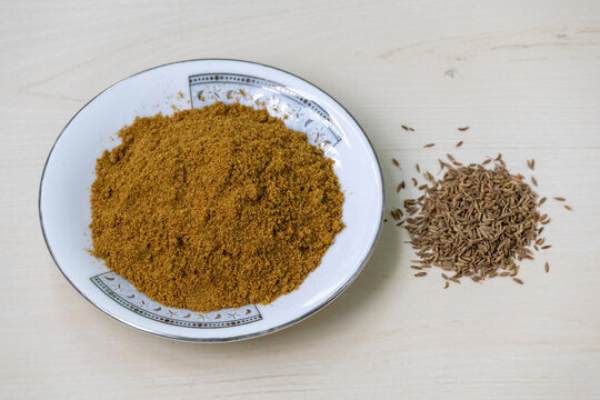 Cumin seed powder is piled on a plate, with a small pile of whole cumin seeds kept beside. Cumin seed powder is a versatile spice with a warm, earthy flavor. It is also known as jeera, jira, or zeera.