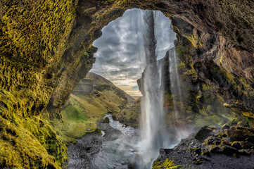 View from behind the Kvernufoss waterfall in Iceland, showcasing a hidden gem with lush green moss, rugged rock formations, and a cascading waterfall in a picturesque landscape.