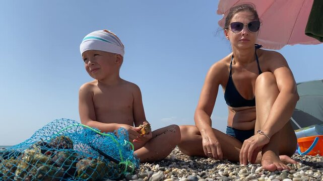 Woman and child look at fresh catch of rapa shells in a net. Mother with little boy enjoying beach holiday on vacation. Evpatoria, Crimea. Rapana venosa