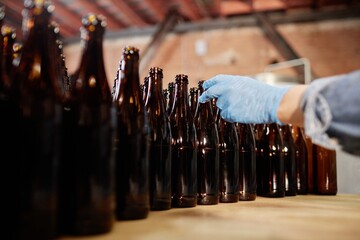 Cropped shot of unrecognizable production worker in protective gloves reaching to take glass bottle...