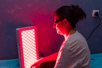 Senior Woman doing the Red Light Therapy Session with Panel and wearing eye protection at home on the bed