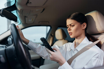 Woman in car driver's seat holding cellphone, glancing at screen, and gripping steering wheel,...