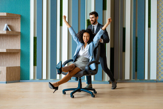 Two young cheerful business people in business wear having fun while racing on office chairs and smiling - Collegues celebrating a successful deal at work, happy and joyful moments in the office