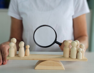 Person holding a magnifying glass over wooden figurines on a balance scale representing focus