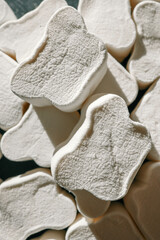 A close-up of fluffy, textured white marshmallows shaped like bones, arranged haphazardly under soft natural lighting. Shadows create depth, highlighting their unique surface details