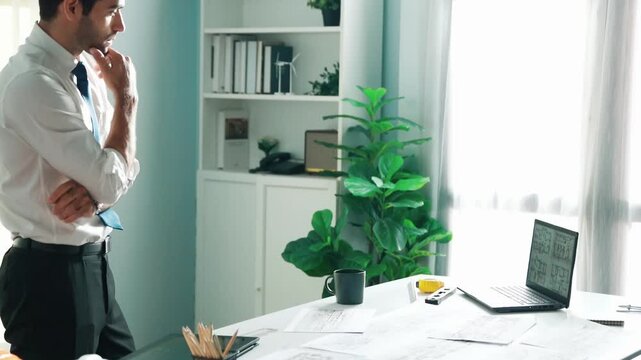 Professional civil engineer standing and drinking coffee and thinking about house plan at meeting table with equipment scatter around. Stressful project manager looking at blueprint. Alimentation.