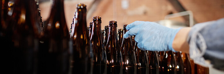 Header image of production workers hand in protective glove reaching to take glass bottle from stack while working at craft beer factory, copy space