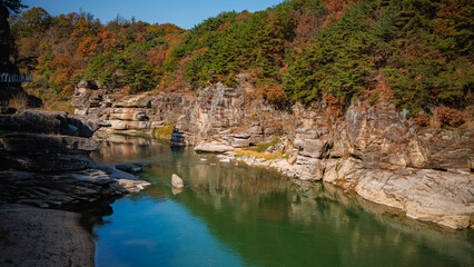 Fototapeta premium Autumn scenery of the Hantan River Columnar Joint Road, with canyons and cliffs formed by the river flowing through the volcanic terrain. (emerald water, blue sky and colorful leaves)